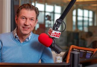 Hake in blue v-neck sweatshirt with a blue plaid collar button up underneath smiling with a WNYC microphone off to his right. Background: Studio