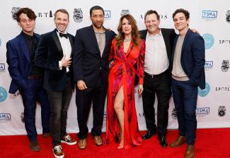Six individuals are arranged in a line on a red carpet. They all wear semi-formal clothing with the fourth person in a bright red dress with black patterns. Behind them is a white step-and-repeat backdrop bearing various logos. 