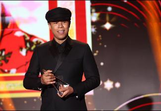 Graise stands onstage with his head turned toward his left. He wears an all-black outfit, including a black cap. He is holding a statuette, a piece of glass shaped like a tall pyramid.