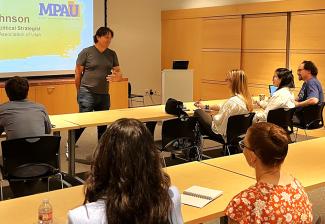 Johnson wears a T-shirt and jeans and stands in front of a group of diverse people seated at tables in a meeting room with a screen displaying the MPAU logo. 