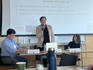 From left, Warren, Powell, and Iovine are positioned around a table. Powell, smiling, stands with a microphone in her hand. She wears a tan blazer and black outfit. Warren looks down at the table. He wears a blue dress shirt and dark slacks. Iovine looks towards Powell. She wears a black shirt. Behind them is a projector screen; in the foreground are some items on a table: a green paper cup, a soda can, and a blue water bottle.