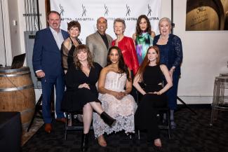 A diverse group of nine individuals in two rows. Three women in the first row are seated; the remaining six stand. There is a SAG-AFTRA SAG Awards step-and-repeat backdrop behind them. 