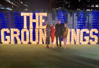 Un plano general de cuatro personas, tres mujeres y un hombre, de pie en una zona cubierta de hierba. Detrás de ellos hay una serie de letras iluminadas que forman la palabra "The Groundlings" en mayúsculas. Se ve el horizonte del centro de Los Ángeles por encima de un muro alto. Se ve algo de iluminación azul.
