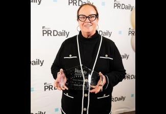 Greenwalt stands on the red carpet before a white step-and-repeat backdrop with the PR Daily logo. She wears a black sweater with white trim and gold buttons and a black turtleneck shirt. In her hands is a glass statuette bearing text in white.