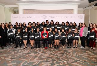 Greenwalt and Reich stand on either side of a large group of young women arranged into two rows. Those in the first row are seated. All the young girls hold their bookbags, black with a thick silver band across the front. Behind the group is a light pink step-and-repeat backdrop with various logos in a darker rust color.