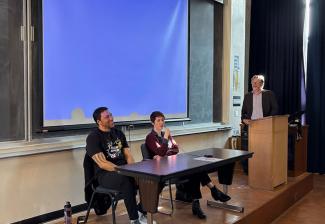 Alton and Elmaleh are seated at a small table in the front of a lecture hall. Dr. Griffey stands to the far right and looks toward the two individuals. Behind them is a projector screen that glows blue.
