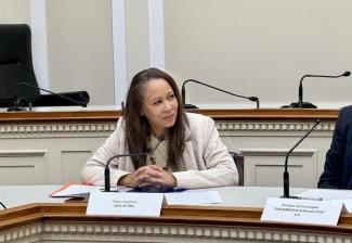 Hutchins is seated at a table in a nondescript room. She wears a cream-colored blazer and bridges her fingers together as she intently looks to her left at an unseen speaker. Positioned before her is a microphone and underneath her hands are papers. Paper name placards for Hutchins and Vanhuynegem are visible. 