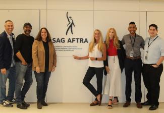 Six individuals stand smiling with the SAG-AFTRA logo (a black figure and its upraised right arm with “SAG-AFTRA” underneath) in the middle.