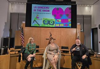 Three individuals sit in directors’ chairs with a colorful presentation behind them on a screen.