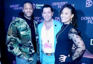 Tres personas están de pie frente a un telón de fondo con el logo del Festival de Cine de Denver. El primer hombre lleva una chaqueta de camuflaje, una sudadera negra con capucha y vaqueros, con las manos en los bolsillos. El segundo hombre lleva un traje azul y una camisa blanca. Alrededor de su cuello lleva un cordón y sus brazos rodean al primer hombre y a la mujer. La mujer de la derecha lleva un vestido negro sobre un vestido negro transparente y algunas joyas.