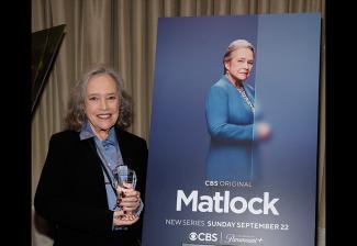 Bates beside a “Matlock” television show poster in a carpeted room with low lighting. Bates wears a black pantsuit and a blue shirt, and holds her awards statuette. The poster is propped on a large easel. 