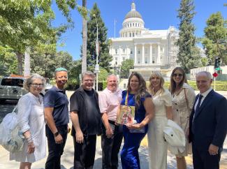 A diverse group of eight people stand outside. In the center right, a woman holds a copy of ‘SAG-AFTRA’ magazine. Behind them is a large white government building.