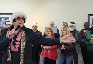 En primer plano, un hombre que imita a Elvis Presley canta con el brazo izquierdo extendido. Lleva una chaqueta negra, una bufanda con estampado de leopardo y un collar de flores rosa, gafas de sol con montura dorada, patillas y un gorro de Papá Noel. Detrás de él, en dos filas, hay un grupo de cantantes. Muchos llevan ropa festiva: rojo o verde y gorros de Papá Noel.