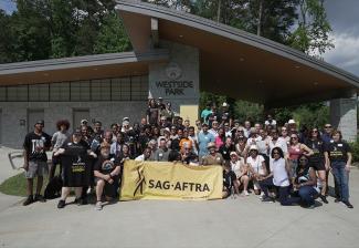 A large, diverse group outside in a park. Many wear black SAG-AFTRA T-shirts. In the center front row, some hold a large yellow banner with the SAG-AFTRA logo.