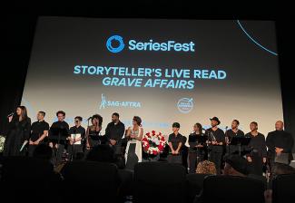 Un grupo numeroso y diverso está en el escenario. Todos los que se ven van vestidos de negro. En el proyector al fondo hay una tarjeta de título con los logotipos y el texto de SAG-AFTRA y SereiesFest, "Storyteller's Live Read 'Grave Affairs'".