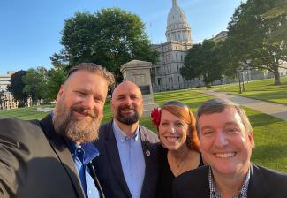A selfie of four individuals, three men and one woman, in a large park area at sundown. Behind them is a large off-white government building.