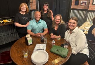 Cinco personas, cuatro mujeres y un hombre, en el interior de un restaurante. El hombre y dos mujeres están sentados a una mesa; las otras mujeres están a ambos lados del hombre. Sobre la mesa hay bebidas, un menú, platos y un bolso verde.