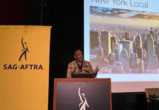 Knight stands behind a podium with the SAG-AFTRA logo. To the side on a projector screen is a bird's eye image of New York City. 