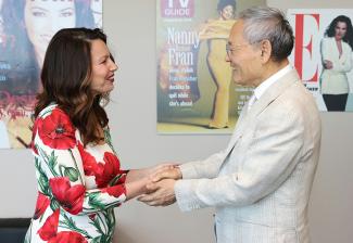 Drescher and Yu In-chon look at each other while grasping each other's hands. Drescher wears a dress with bright red flowers; Yu wears a light colored blazer and white dress shirt. Behind them are various magazine posters bearing Drescher's face.