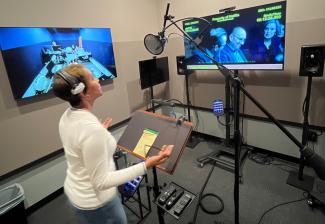 A woman stands in a recording studio with equipment, reciting lines as a film plays on two screen. 