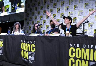 Burch, Woodward, Dalal y Elmaleh están sentados en una mesa con manteles con el logo de la Comic Con de San Diego. Elmaleh se inclina hacia adelante en su asiento con los brazos abiertos mientras los demás observan.