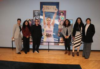 Six individuals stand in front of a painted collage. They are grouped in two sets of three and there is a large gap between them. Visible is a painting of a person holding a sign that reads "Union" in all-cap lettering.