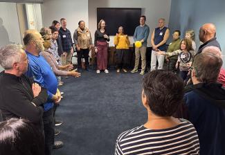Un grupo grande de personas diversas se reúne en círculo en la sala de conferencias del sindicato SAG-AFTRA de Nueva Inglaterra; un miembro sostiene una pelota amarilla mientras se prepara para un ejercicio de actuación.