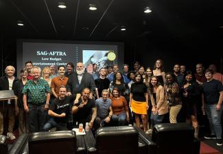 Un grupo grande de unas 30 personas posa junto a un teatro con poca luz, sonriendo para la foto. Detrás de ellos, una pantalla muestra una presentación titulada "Contratos de entretenimiento de bajo presupuesto de SAG-AFTRA". El grupo incluye hombres y mujeres de diversas edades y etnias, algunos con ropa informal y otros con atuendos de negocios.
