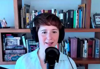 Elmaleh is seated at an unseen table. She wears a white shirt with buttons. In front of her is a recording mic; she also wears headphones. Behind her on shelves are various books and games.