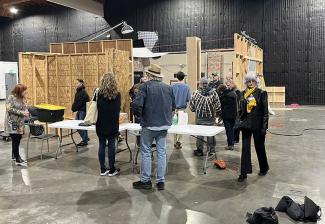 Personas vestidas con ropa informal se arremolinan en un gran espacio que parece un almacén. Hay mesas de plástico y, en una parte de la sala, hay paredes de madera independientes que forman un decorado.