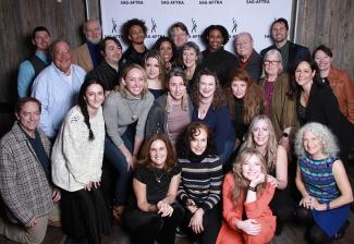 Un grupo de aproximadamente 30 personas sonrientes, algunas de pie y otras arrodilladas, se reúnen frente a una pancarta de SAG-AFTRA con pasos y repeticiones.