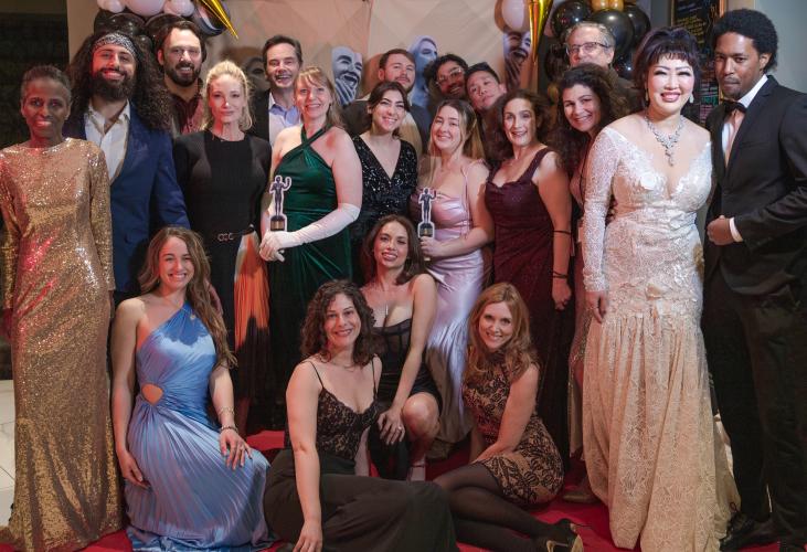 A couple dozen diverse individuals take a group photo. In the center front, three women are seated on the red carpet; one woman to the left kneels. All dressed in formal attire. Behind them is a step-and-repeat backdrop with balloons and award posters.