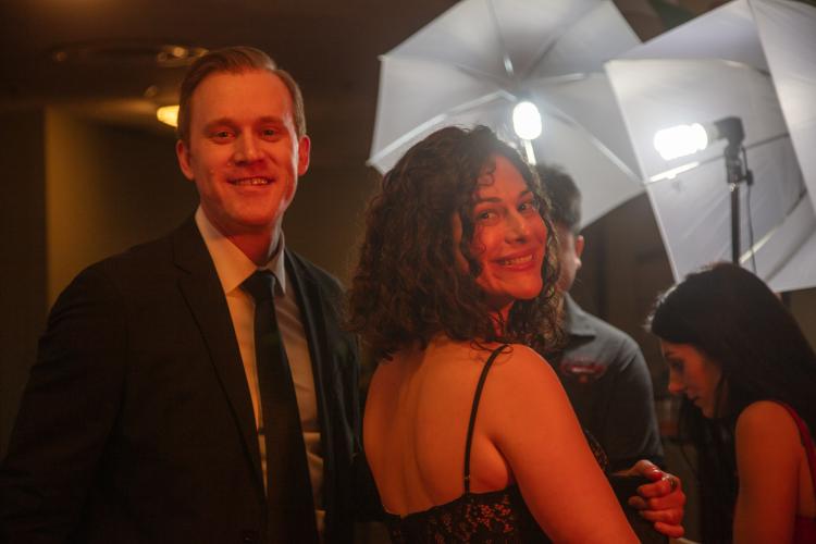 Rust and Speert look towards the camera, with Speet looking over her right shoulder. Rust wears a suit and tie; Speert wears a black dress with thin spaghetti straps. A red light shines on their faces.