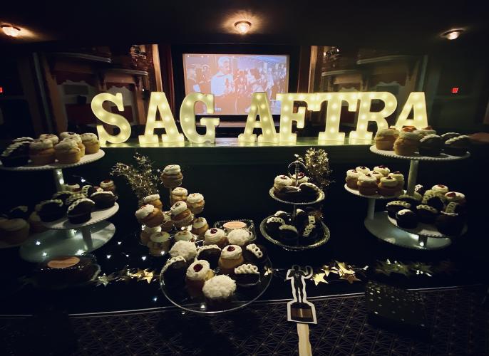 A stylized image of different cupcakes being displayed. The cupcakes are arranged on flat and multi-tiered trays; there is also a paper cut-out of the award statuette. Above are brightly-lit letters arranged to spell “SAG-AFTRA.”