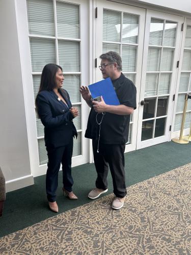 Ortega y Astin son capturados en medio de una discusión. Ortega viste un traje pantalón azul oscuro; Astin está vestido completamente de negro y sostiene su teléfono, auriculares y una carpeta azul. Detrás de ellas hay puertas con persianas cerradas.