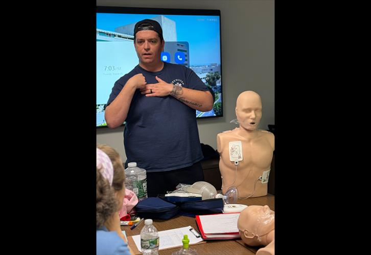 Un hombre con una gorra de béisbol al revés, una camiseta azul y pantalones color caqui se encuentra de pie frente a un aula. Muestra cómo utilizar un desfibrilador externo automático. Hay un maniquí adulto de reanimación cardiopulmonar colocado sobre una mesa.