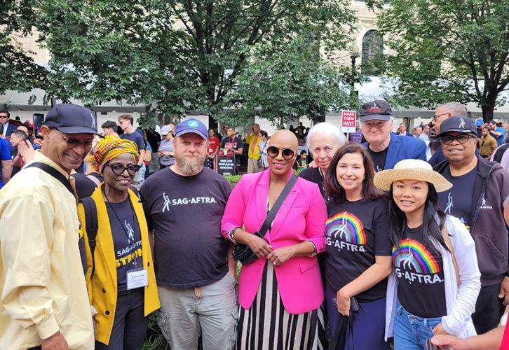 Un grupo diverso de personas se encuentra de pie a ambos lados de la representante Ayanna Pressley, en el centro, en un área al aire libre. Pressley lleva un vestido de rayas blancas y negras con un blazer rosa y una cartera negra cruzada. Tres personas se encuentran de pie a un lado de ella y cinco personas se encuentran del otro lado, todas ellas con una variedad de camisetas de SAG-AFTRA.