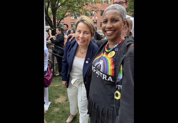 Healey viste pantalones blancos, una blusa blanca y un blazer azul, y está junto a Lyman, quien viste una camiseta negra SAG-AFTRA Pride y un collar de arcoíris.
