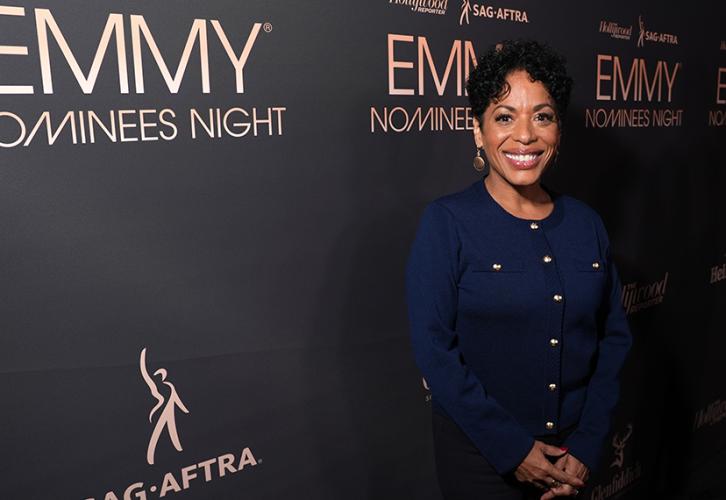 Colón-Zaya is oriented towards the far right of the photo. Smiling, she wears a blue dress with brass buttons down its length and breast pockets. Her hands are clasped in front of her. Behind her is a step-and-repeat backdrop, black with the THR/SAG-AFTRA Emmy Nominee Night party logo in gold.