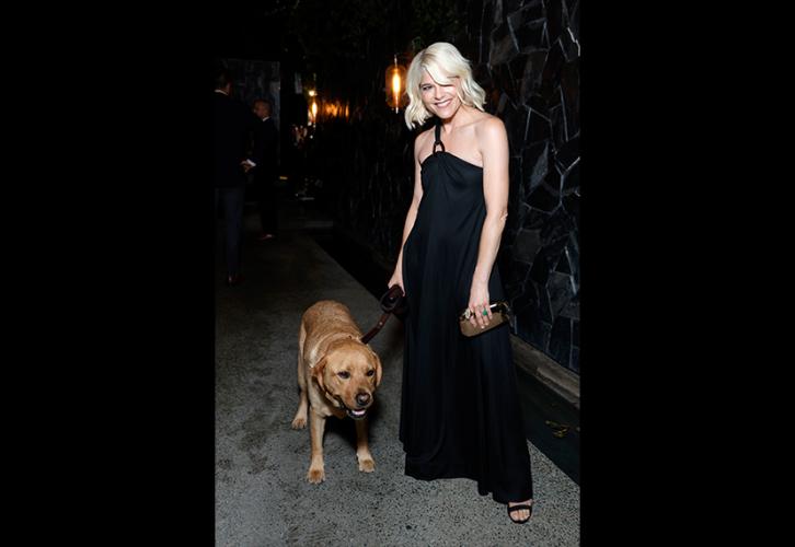 Blair stands outside in a courtyard. She wears a black maxi dress and holds a small purse in her left hand; in her right hand is a dog leash. On her right is a large dog.