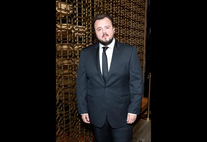 Bradley wears a black suit, a white dress shirt and black tie. He stands with his hands at his side. Behind him is a wall patterned with geometric shapes in gold.