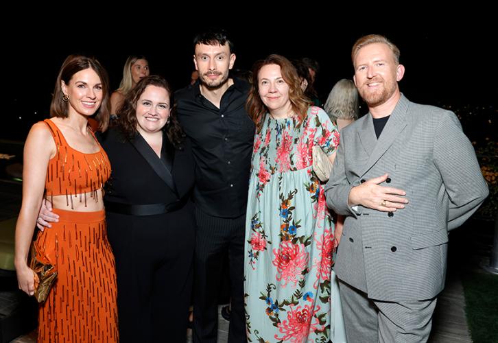Raine, Gunning, Gadd, the guest and Goodman-Hill stand closely together. Raine wears an orange two-piece top and skirt set with tassels; Gunning a black dress; Gadd, an all-black outfit with his guest in a pale-blue dress patterned with pink flowers; and Goodman-Hill in a gray suit with a black shirt. Goodman-Hill’s hand is over his chest. Behind the group are party minglers.