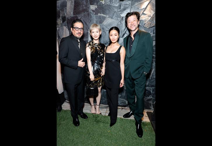 Sanada, Hoshi, Sawai and Asano stand closely together. Sanada wears a black suit with a distinctive tie; Sanada a dark-gold metallic dress and bag; Hoshi, a black dress; and Asano, a dark-green suit with a black dress shirt. Behind them is a wall with gray slates.