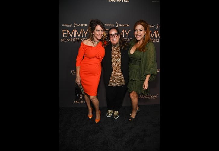 From left, Fisher, Greenwalt and Walter stand closely together. Fisher’s dress and shoes are orange; Greenwalt wears a black pantsuit, leopard-print shirt and silver sneakers; and Walter, a green dress. Behind them is a step-and-repeat backdrop, black with the THR/SAG-AFTRA Emmy Nominee Night party logo in gold.