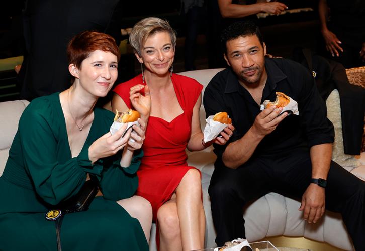 Elmaleh, Norris and Alton are seated on a white couch, each holding a burger. Elmaleh wears a long green dress; Norris a red dress; and Alton an all-black outfit. Elmaleh and Alton lean forward.