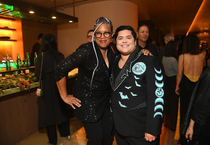 Gipson and Guillén stand near each other adjacent to a bar area. Gipson wears a black sequined top and black pants; Guillén, a black blazer with light blue bats and phases of the moon. Gipson’s arm is around Guillén’s shoulders. Behind them are party attendees.