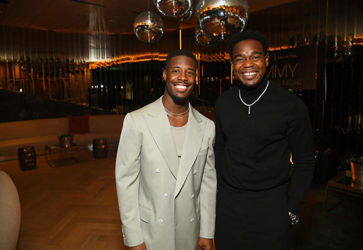 Walker and Darden stand closely together in what appears to be a dance area with disco balls overhead. Walker wears an all-gray ensemble and thin neck chain; Darden, an all-black outfit with a silver chain. Both men smile widely. 