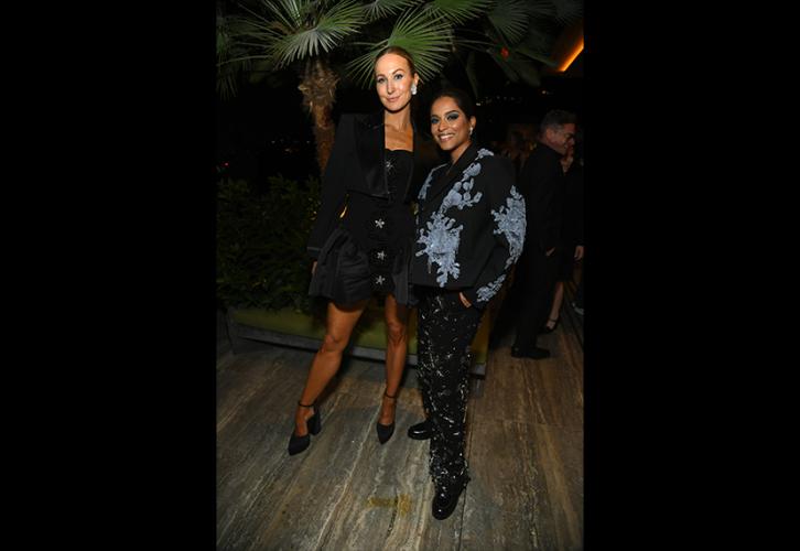 Glaser and Koshy stand outside in a courtyard. Glaser wears a black dress with buttons down its length and a cropped black blazer; Koshy wears a black blazer with a blue pattern and sequined black pants. Glaser poses with her right leg extended; Koshy’s hands are in her pockets. Behind them is a palm tree.