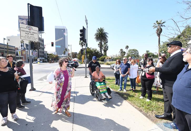 A wide shot of a scene on a street. Ginsburg Bikel, wearing a flowered kimono robe over a fuschia dress, holds a microphone. The crowd frames itself around her. Visible in the photo are Tina Calderon, center background, and Dan Navarro, far right. The Theodore Bikel Square sign is visible in the background, far left.