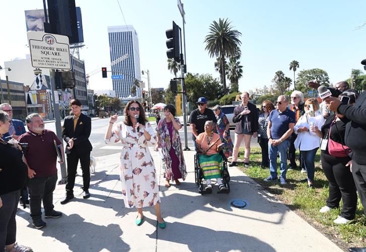 Yaroslavsky, wearing a patterned dress speaks into a microphone. The crowd frames itself around her. Visible in the photo are Aimee Ginsburg Bikel and Tina Calderon, center background. The Theodore Bikel Square sign is visible in the background, far left.